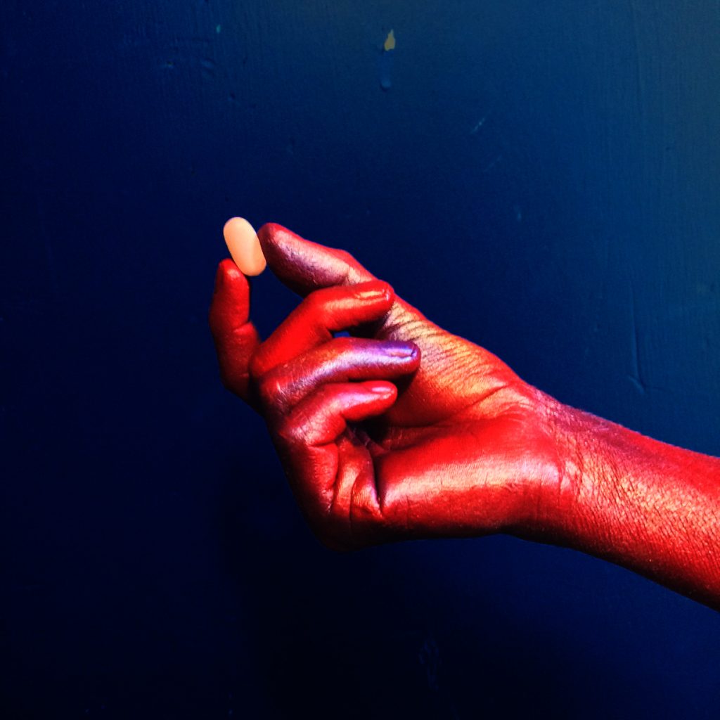 An image still from Jorge Bordello’s “Ministry of Health” shows a hand painted with red body paint holding an orange pill between the index finger and thumb